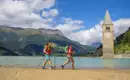 Zwei Wanderer am Ufer des Reschensees in Südtirol mit Kirchturm im Wasser.