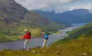 Zwei Personen wandern auf einem Hügel mit Blick auf Loch und Berge in Schottland.