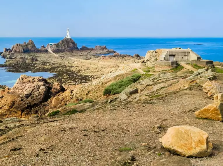 Ein Leuchtturm auf einer felsigen Klippe vor dem Meer mit blauem Himmel.