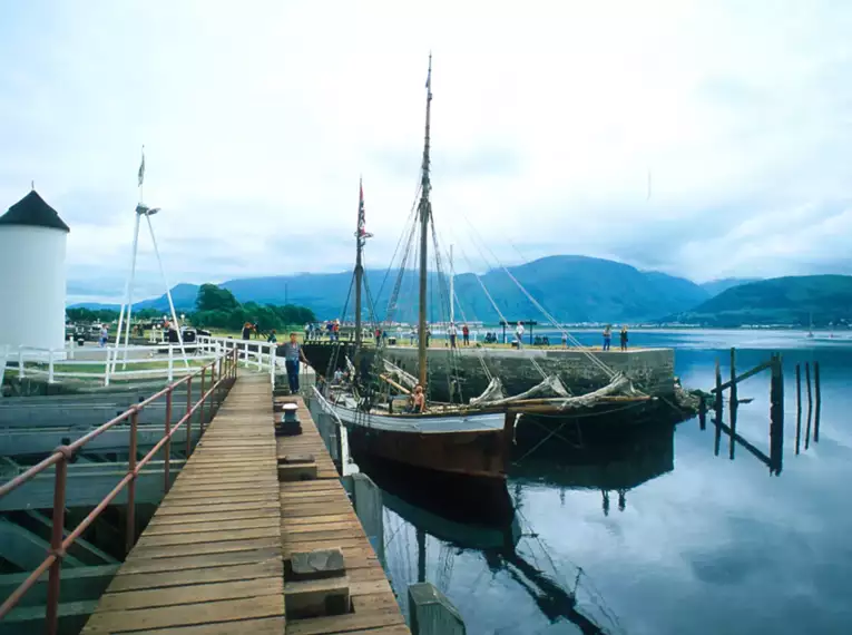 Ein traditionelles Segelschiff liegt im Hafen von Fort William, Schottland, vor Anker. Im Hintergrund sind Berge zu sehen.