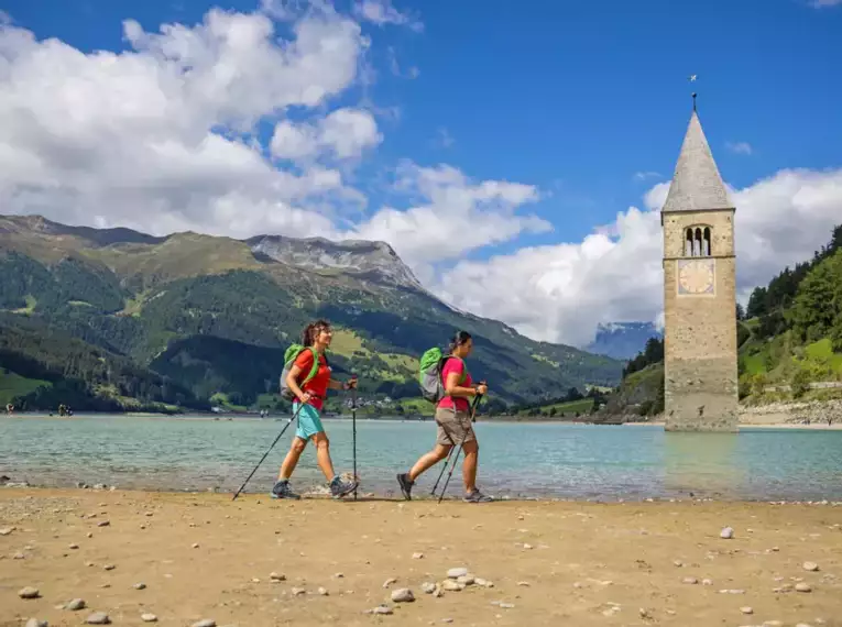 Zwei Wanderer am Ufer des Reschensees in Südtirol mit Kirchturm im Wasser.