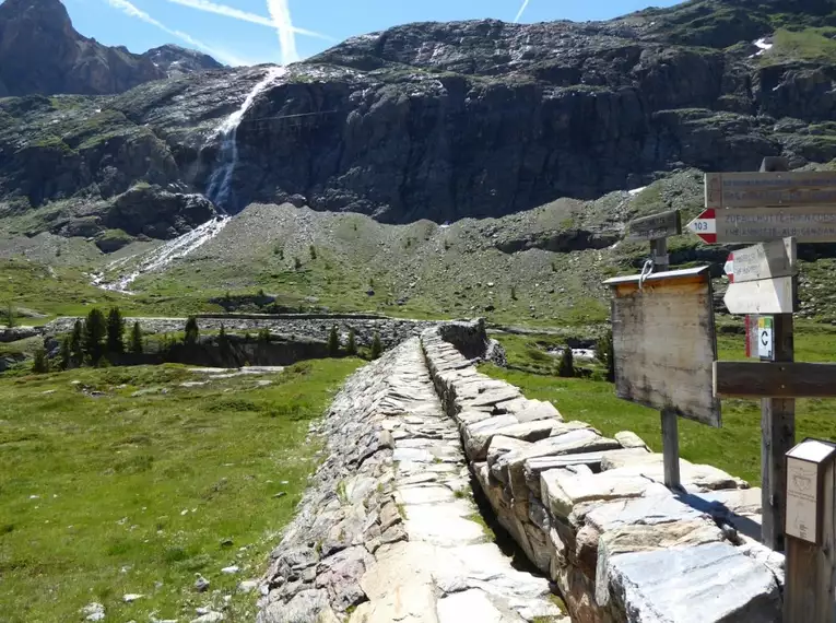 Steinpfad mit Wegweisern, Bergkulisse und Wasserfall in Südtirol.