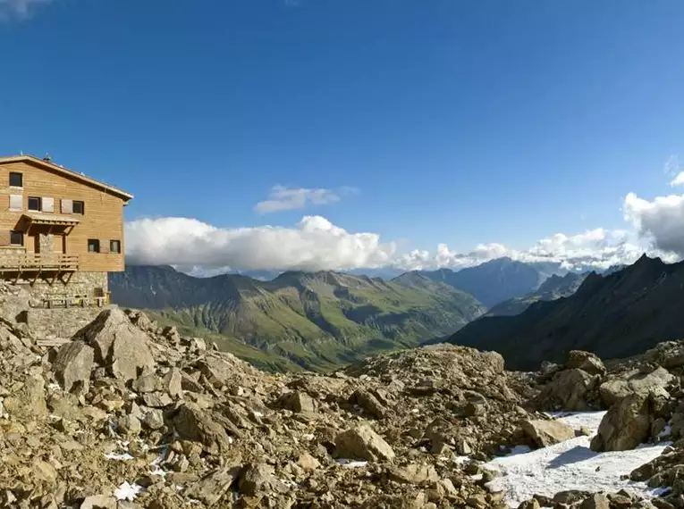 Alpenhütte umgeben von majestätischen Berggipfeln und sonniger Landschaft.