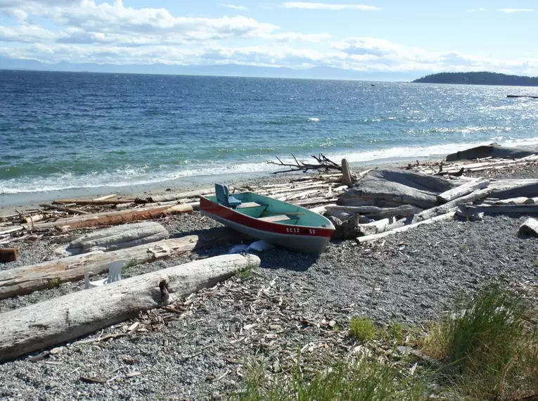 Ein kleines Boot liegt auf einem Kiesstrand mit Treibholz, dahinter das weite Meer von Vancouver Island.