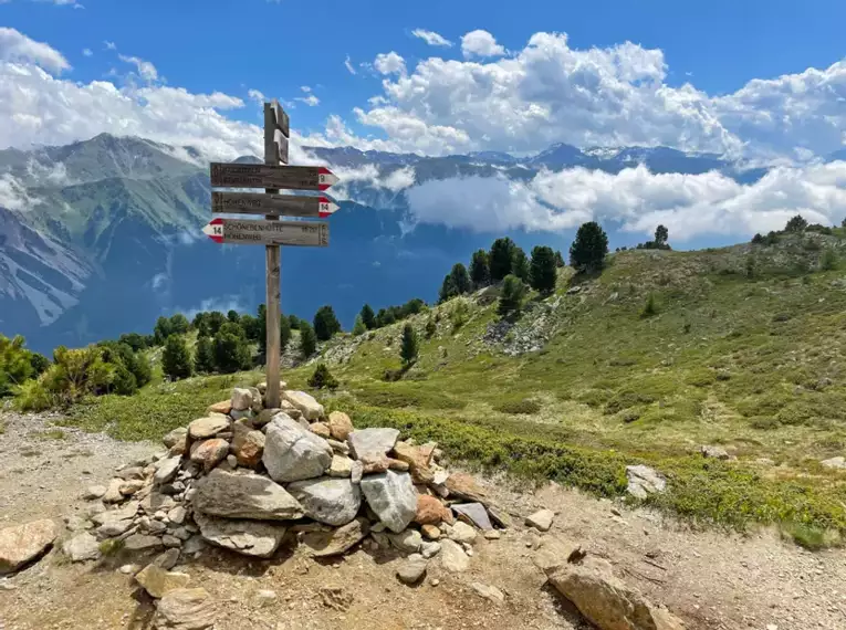 Holzwegweiser auf einem alpinen Wanderpfad mit Berglandschaft im Hintergrund.