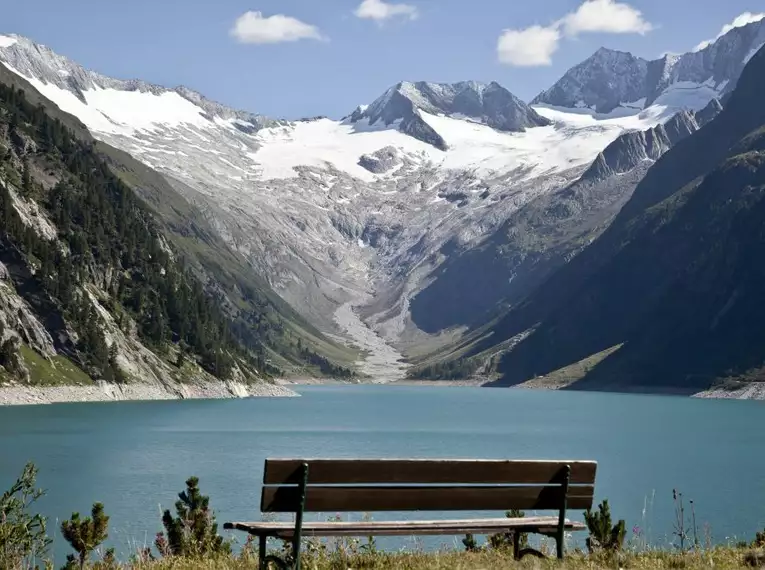 Holzbank vor einem See in den Alpen mit schneebedeckten Bergen im Hintergrund.