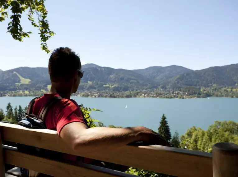 Person sitzt auf einer Bank mit Blick auf den Tegernsee und umliegende Berge.