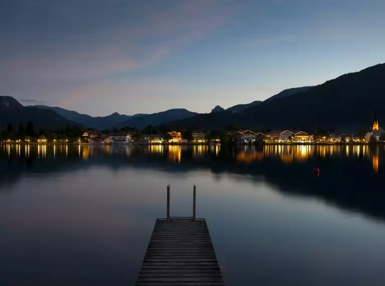 Nächtliche Ansicht von Tegernsee mit Lichtern und Steg am Wasser.