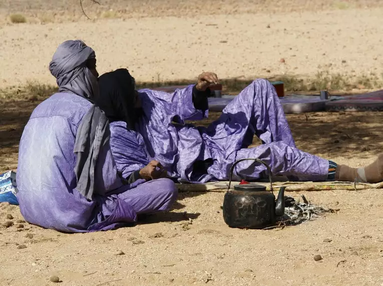 Zwei Personen in traditionellen Gewändern ruhen im Schatten in der Sahara.