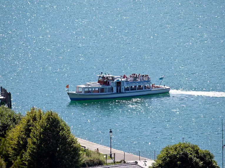 Ein Boot fährt bei Tag über den Tegernsee in Bayern, umgeben von blauem Wasser.