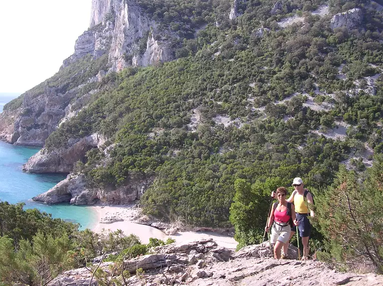 Zwei Wanderer auf einem felsigen Küstenpfad mit Blick auf das Meer und die rauen Felsen von Sardinien.