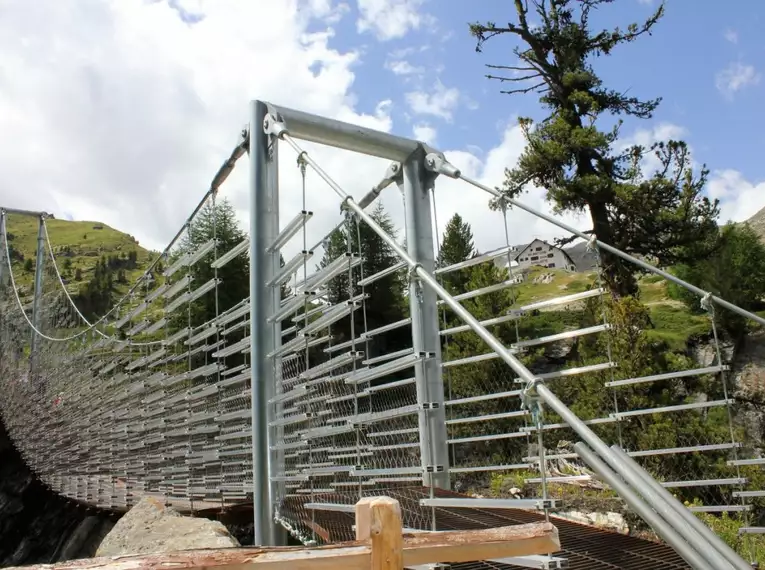 Hängebrücke im Martelltal, umgeben von Bergen und Bäumen, blauer Himmel im Hintergrund.