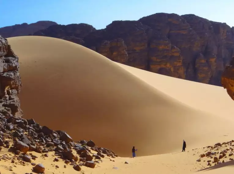 Zwei Personen erkunden die Sahara zwischen Felsen und Sanddünen.