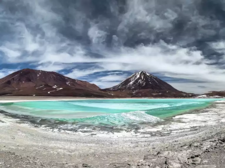 Bizarre Landschaft mit grünblauer Lagune in einer Wüstenregion. Im Hintergrund erheben sich hohe Berge unter dramatischem Himmel.