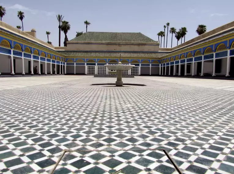 Ein weitläufiger mosaikverzierter Innenhof des Bahia-Palastes in Marrakesch mit umgebenden Gebäuden und Palmen.