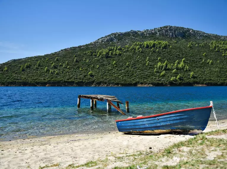 Verlassener Strand mit Boot und Steg, umgeben von grünen Hügeln.