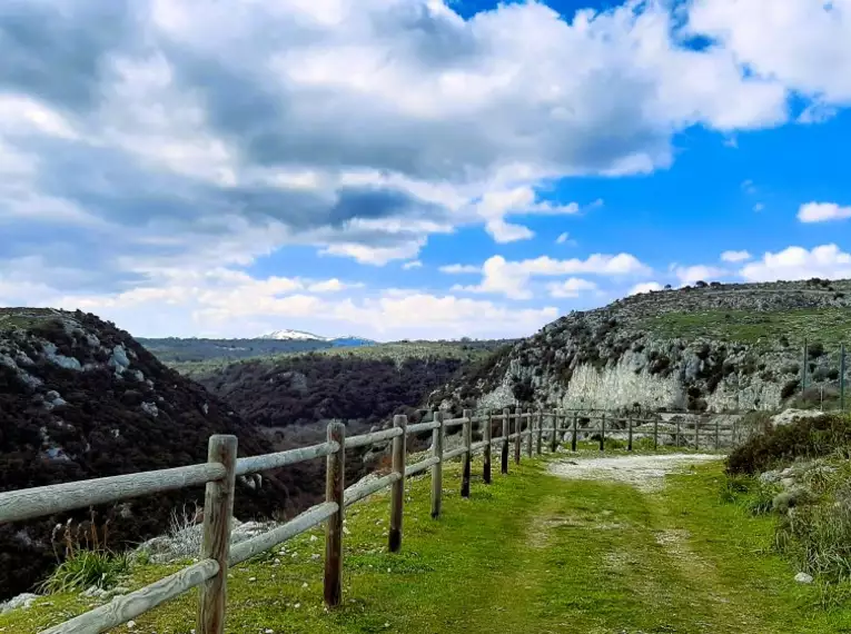 Wanderweg mit hölzernem Geländer in einer grünen, hügeligen Landschaft unter blauem Himmel.