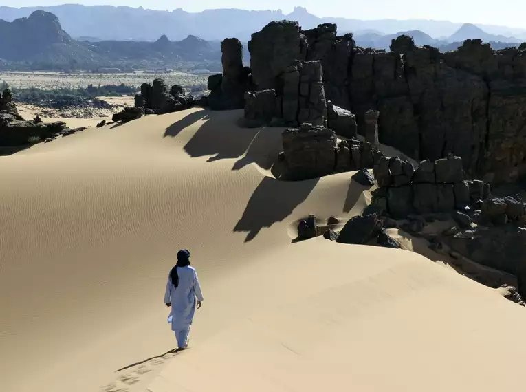 Person in traditioneller Kleidung wandert durch die Sahara mit Felsen im Hintergrund.