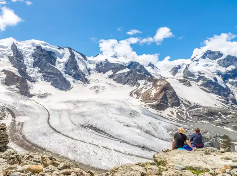 Zwei Wanderer betrachten majestätische Gletscher in den Alpen, umgeben von schneebedeckten Bergen und klarem Himmel.