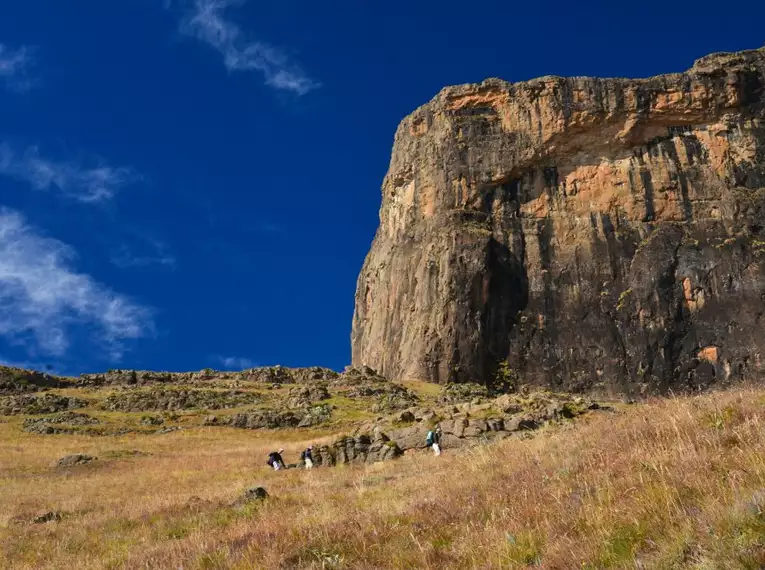 Menschen wandern entlang einer steilen Felswand in den Drakensbergen, Südafrika, unter blauem Himmel.