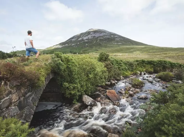 Ein Wanderer betrachtet die beeindruckende irische Landschaft mit einem Fluss und einem bergigen Hintergrund.