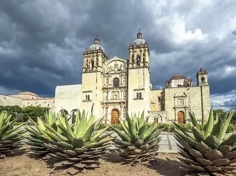 Historische Kirche in Oaxaca, umgeben von Kakteen unter dramatischem Himmel.