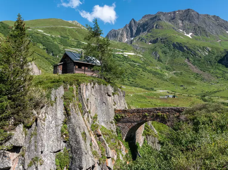 Alpine Landschaft mit Bergen, grünen Wiesen und einer historischen Steinbrücke.