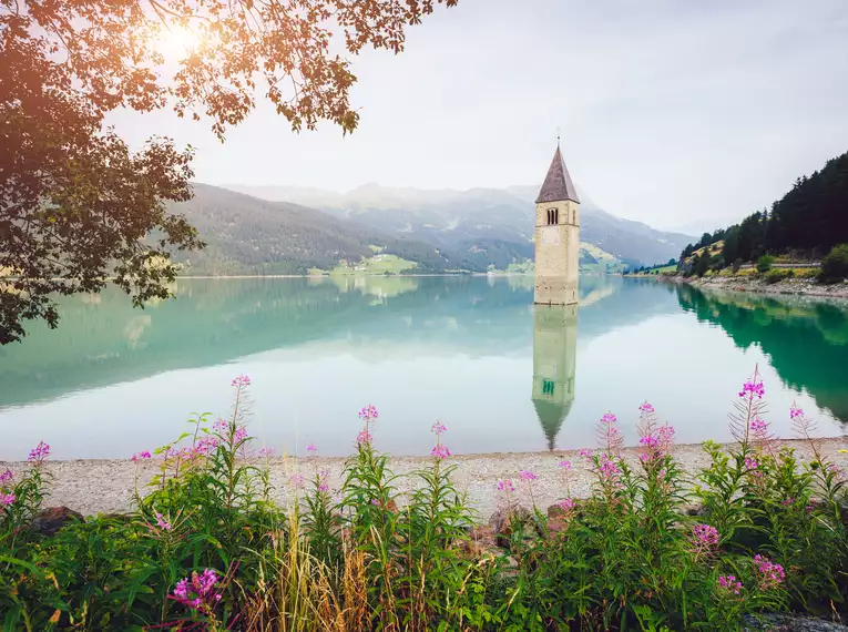 Ein versunkener Kirchturm ragt aus dem Reschensee, umgeben von Berglandschaft.