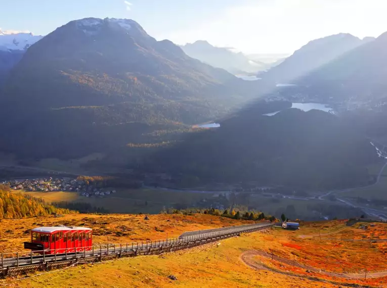 Eine rote Bergbahn fährt durch das herbstliche Tal im Oberengadin.