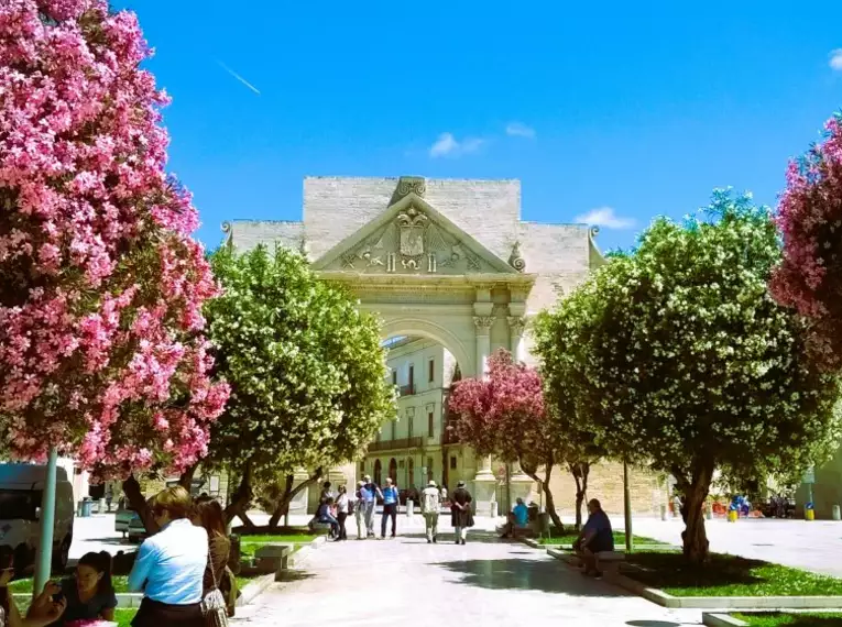 Sonniger Platz mit blühenden Bäumen und altem Gebäude, umgeben von Menschen.