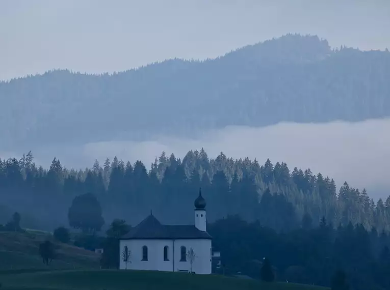 Eine Kirche mit Zwiebelturm vor nebeligen, bewaldeten Alpenlandschaft.